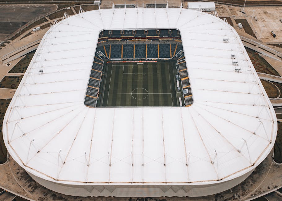 An aerial view of The Hive Stadium showing a large, oval-shaped sports arena with a partially transparent roof supported by white pylons and cables. Inside, the bright green football pitch is clearly